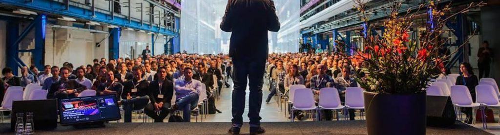 A speaker addressing a full room from behind a lectern, seen from the back of the hall